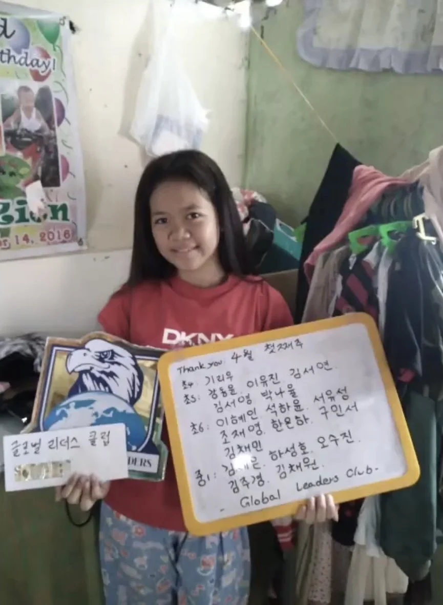A young girl standing indoors holding a framed drawing of an eagle and a whiteboard with handwritten notes in Korean and English. There is a poster on the wall in the background, and clothes hanging nearby.
