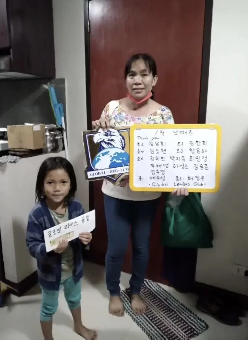 A woman and a young girl standing indoors, holding signs. The woman is holding a sign with a bird and globe logo and a handwritten message in Korean. The girl is holding a smaller sign also with Korean writing. They are near a red door, with kitchen 