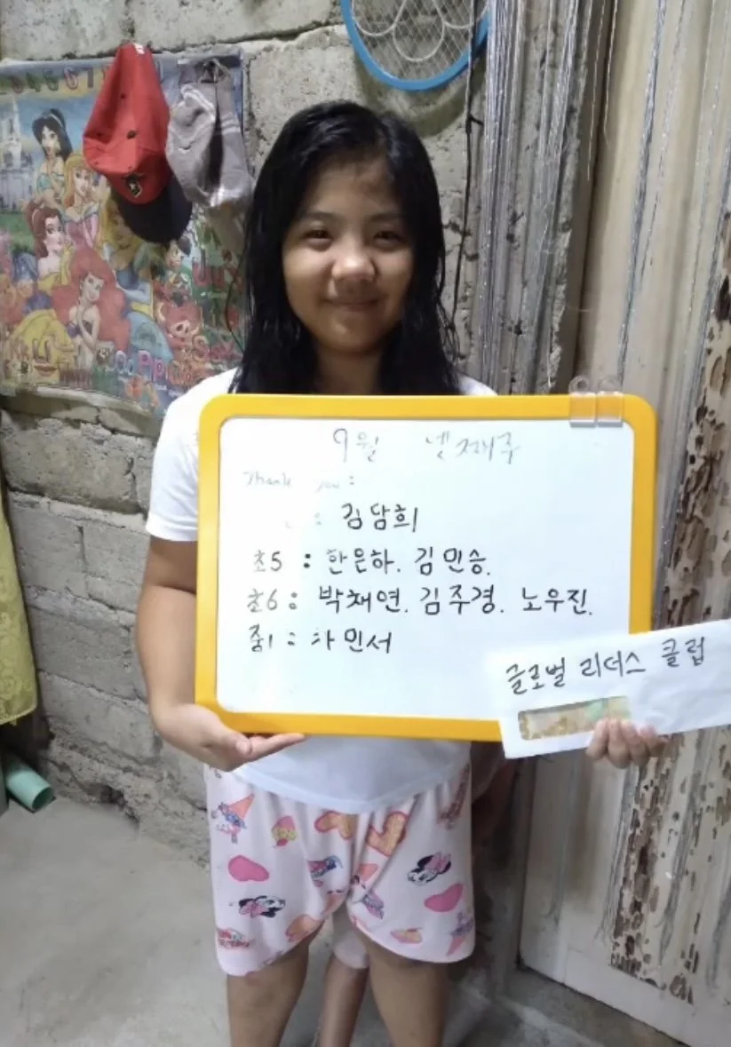 A young girl with wet hair standing indoors, holding a whiteboard with writing in Korean, smiling, with a cardboard wall, a colorful poster featuring cartoon characters, and some hanging clothes behind her.