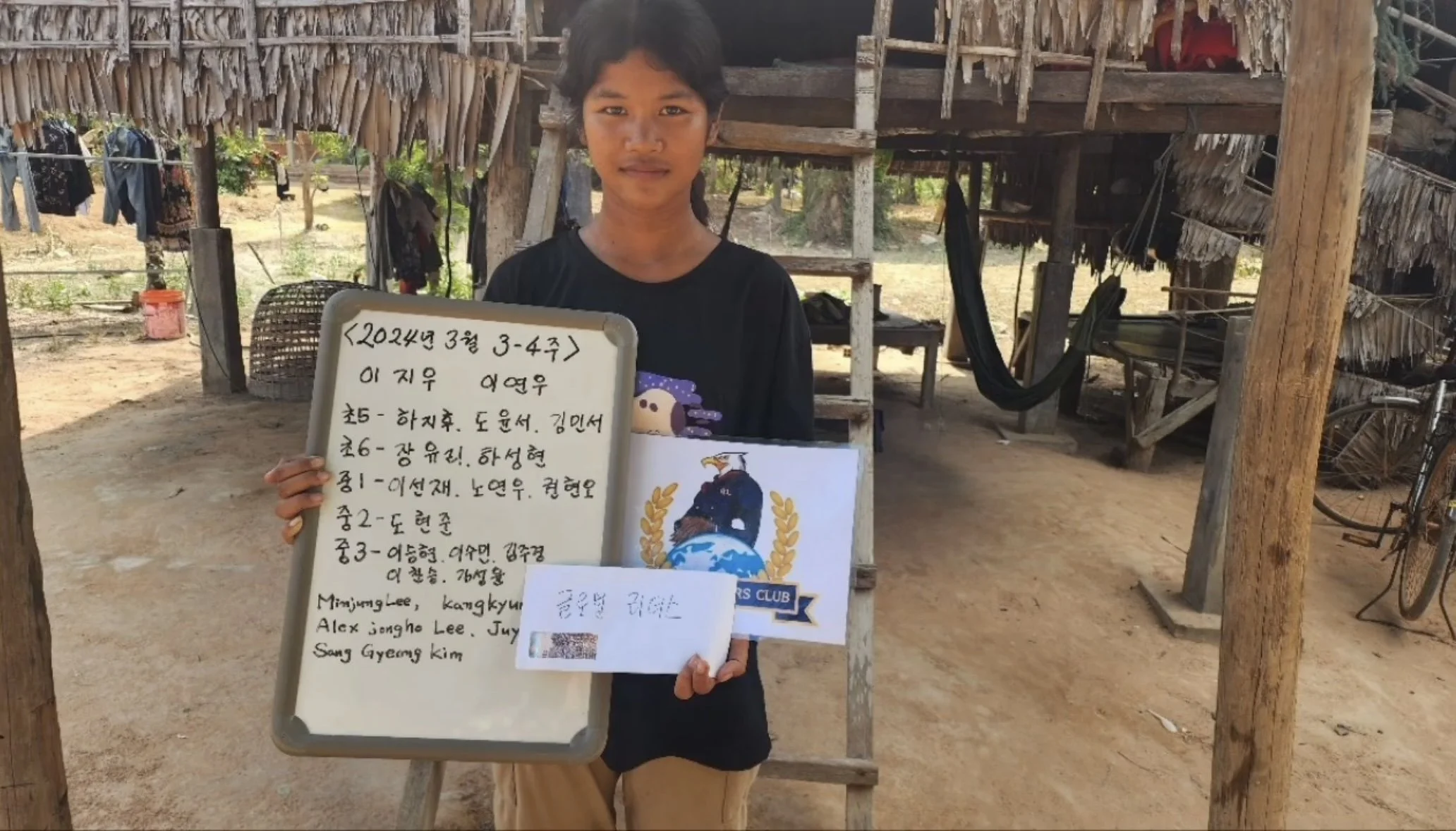 A girl standing outside a rustic wooden structure, holding a whiteboard with writing in Korean and English, and a paper with additional text. The background shows a dirt ground, some greenery, a bicycle, and laundry hanging on a line.