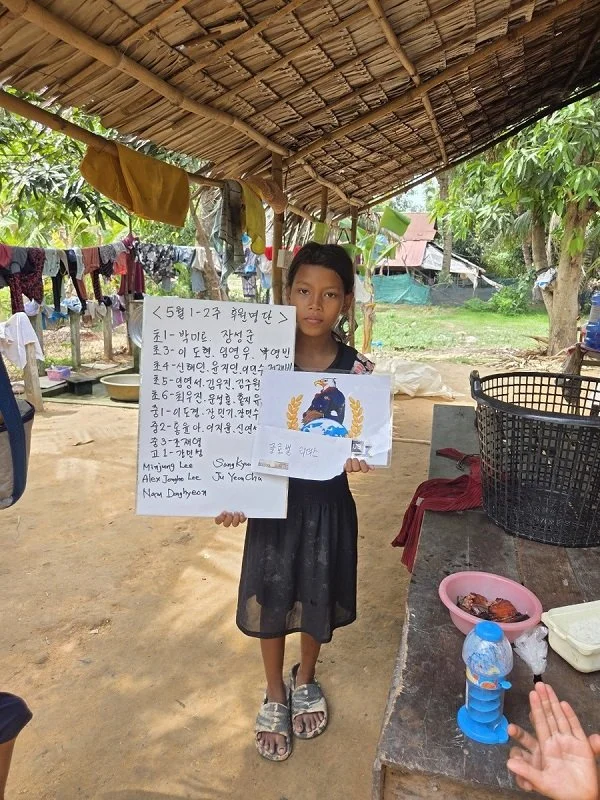 Young girl standing outdoors under a thatched roof structure, holding handwritten notes and a drawing. Clothes hang on a line nearby, and there are trees, a pink bowl with food, and a water bottle on a table.