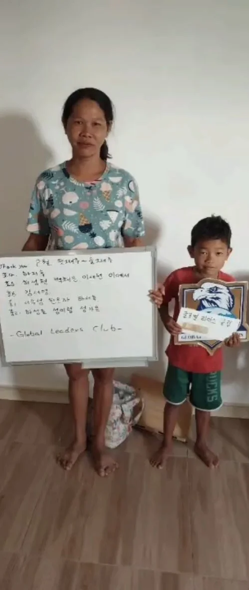 A woman and a young boy standing indoors against a plain wall. The woman is holding a whiteboard with handwritten text, and the boy is holding a shield-shaped sign with an eagle and Korean writing. The woman wears a colorful patterned shirt and the b