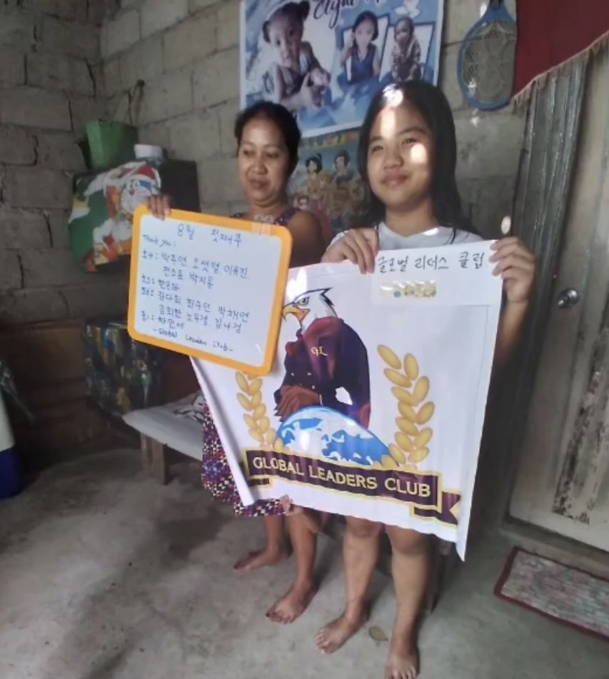 A woman and a girl holding a 'Global Leaders Club' banner inside a room with brick walls. The girl is smiling and holding a whiteboard with handwritten text, while the woman stands behind her. There are posters and decorations on the wall.