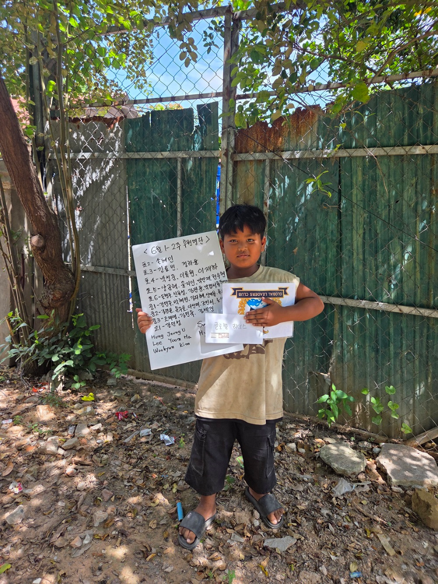 A young boy standing outdoors on a dirt ground, holding posters with writing in Korean and English. The background has a green metal fence, some leafy trees, and blue sky.