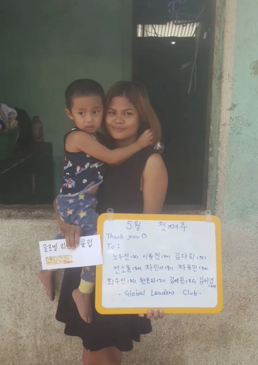 A woman holding a young boy in her arms, standing in front of a wall with a small window and a door in the background. The woman is smiling, and the boy is looking at the camera with a neutral expression. She is holding a whiteboard with handwritten 