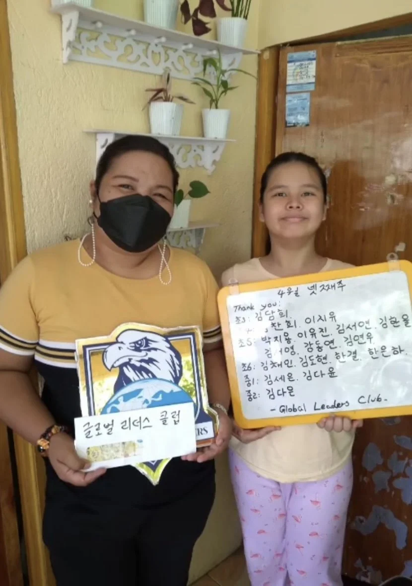 A woman wearing a black mask and a yellow shirt, and a young girl in pajamas, are standing in front of a yellow wall with white shelves holding potted plants. The woman is holding a sign with a logo and Korean text, and the girl is holding a whiteboa