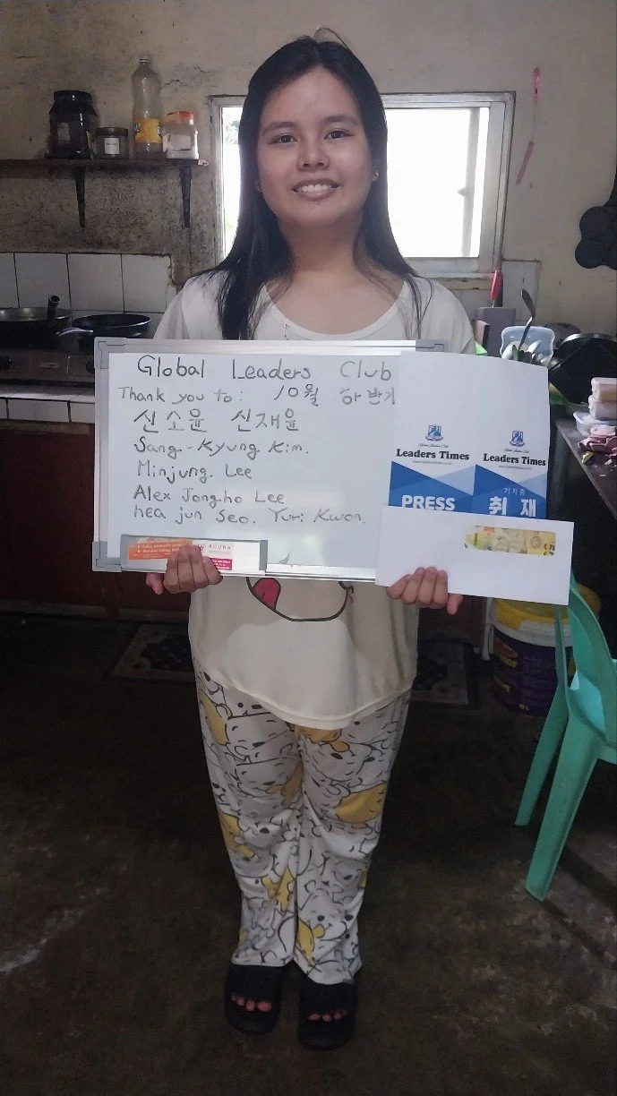 A young girl standing in a kitchen holding a whiteboard with a thank you message and names written on it. She is wearing a white T-shirt, cartoon-printed pajamas, and black slippers. The kitchen has a window, shelves, and various kitchen items in the