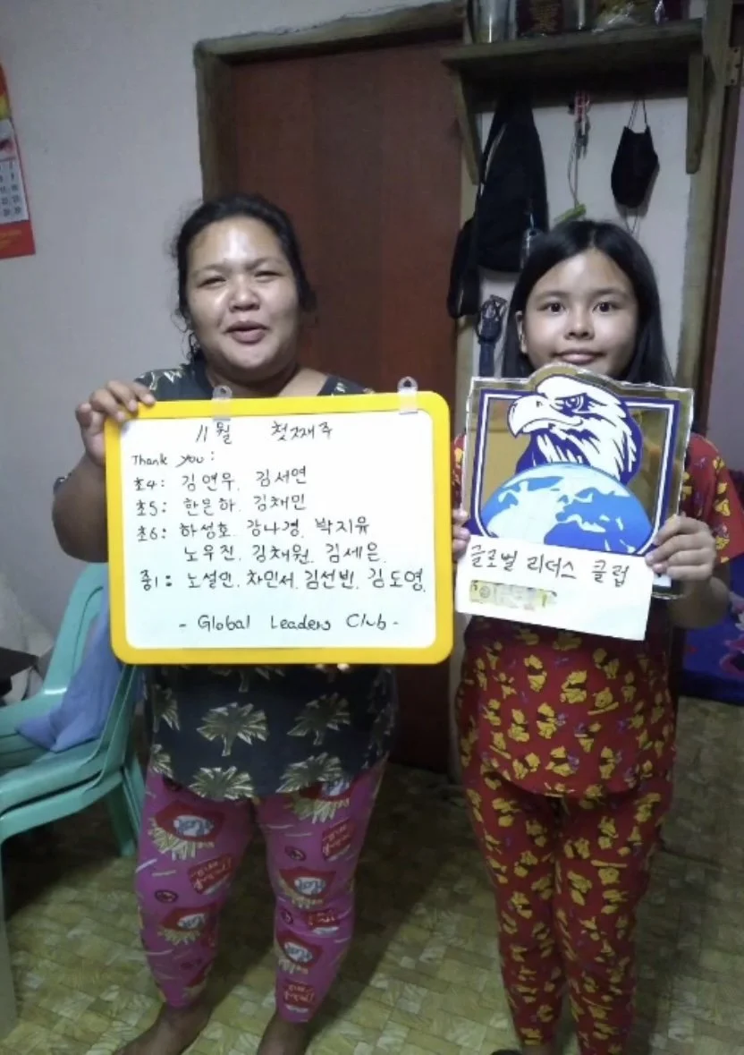 Two women standing indoors, holding signs that feature English and Korean text. The woman on the right is holding a sign with an eagle and globe graphic, and wearing red pajamas with yellow patterns. The woman on the left is holding a whiteboard with