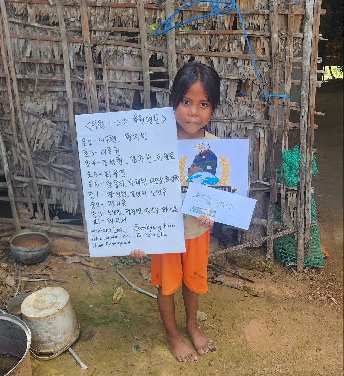 Young girl standing barefoot outdoors, holding handwritten notes and a colorful paper, in front of a bamboo fence with metal buckets on the ground.