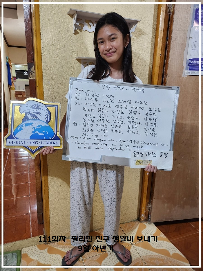 A young woman is standing indoors holding a whiteboard with handwritten text in Korean and some English. She is smiling and wearing a white shirt and patterned pants. There is a badge or sign with a globe and eagle logo reading "GLOBAL 2005 LEADERS" 