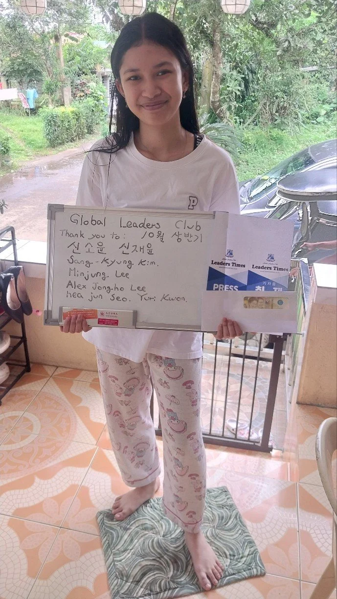 A young girl standing on a fabric on the floor, holding a whiteboard and papers with handwritten notes, indoors with green trees visible through a window behind her.