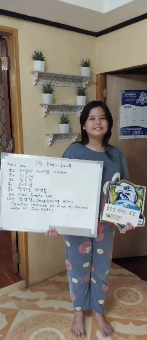 A young girl standing barefoot in a room, holding a whiteboard with handwritten notes and a colorful certificate with a lion logo. The room has a beige wall with three small potted plants on white shelves and a door with a calendar hanging beside it.