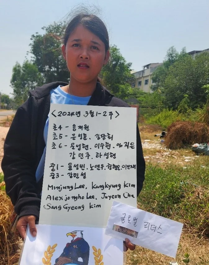 Person holding a sign with multilingual text, standing outdoors with trees and buildings in the background.