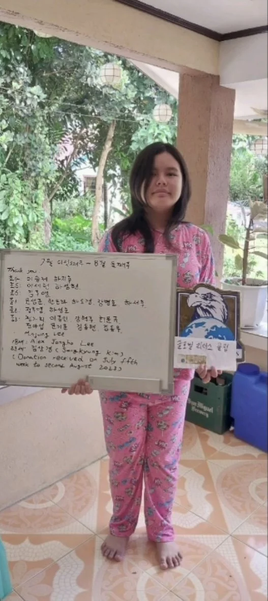 A young girl in pink pajamas holding a whiteboard with handwritten notes and a sign with an eagle and globe. She is standing indoors near a large window with a view of trees and greenery outside.