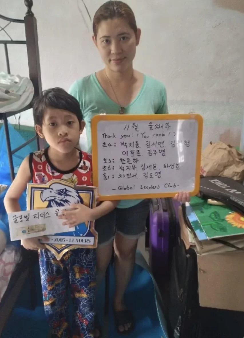 A woman and a young boy standing indoors holding signs and a certificate. The woman has short hair and is wearing a light blue shirt, while the boy has short dark hair, and is wearing a sleeveless shirt and colorful pants. The boy is holding a certif