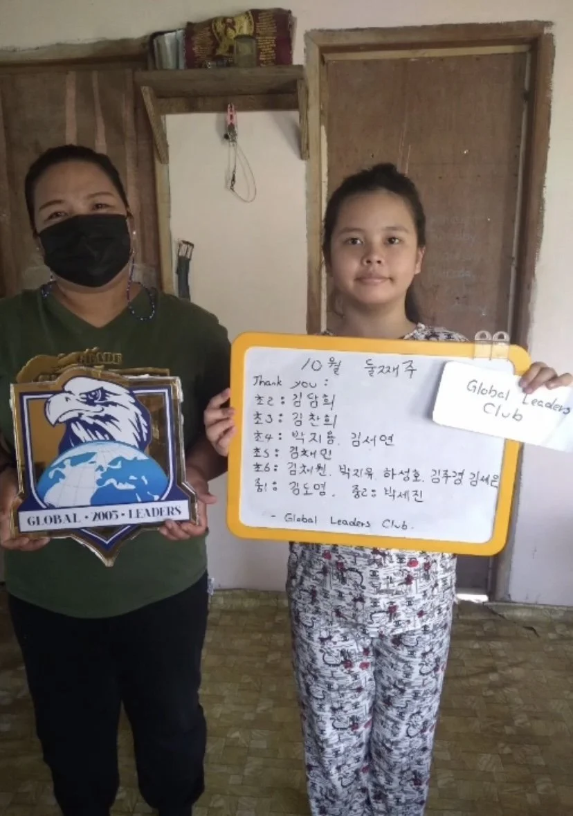 A woman with a black face mask and a girl holding a whiteboard with handwritten notes, standing inside a room with wooden doors and a simple wooden shelf.