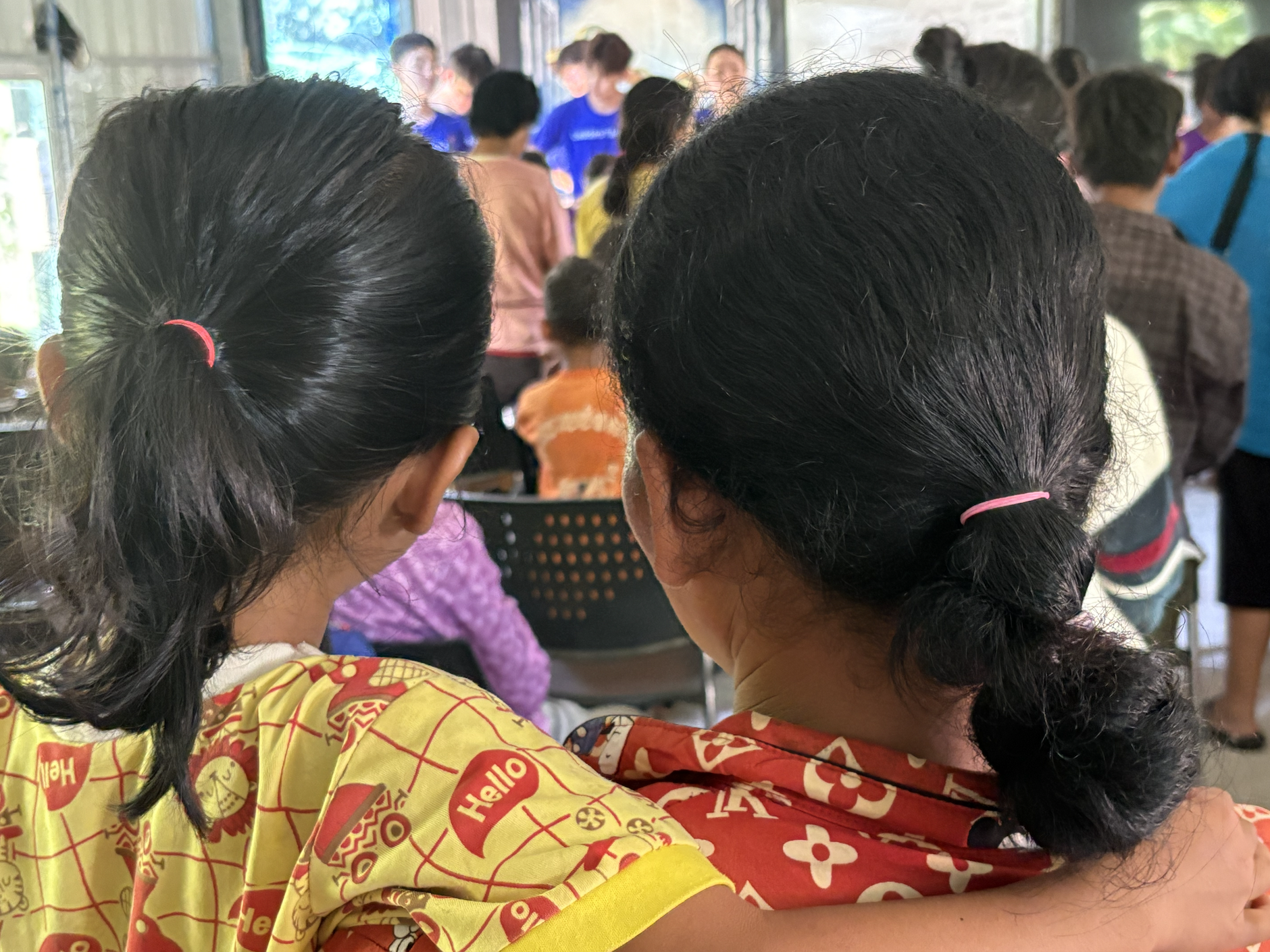 Two women with black hair tied back in ponytails, wearing colorful patterned shirts, are sitting and embracing in a crowded indoor setting with many people standing and sitting around.