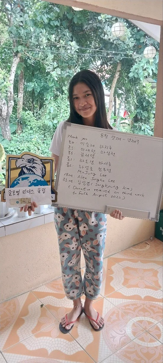 Young woman standing indoors holding a whiteboard with written notes, wearing pajama pants with rabbits, pink flip-flops, and a white t-shirt, with outdoor greenery visible through a window behind her.