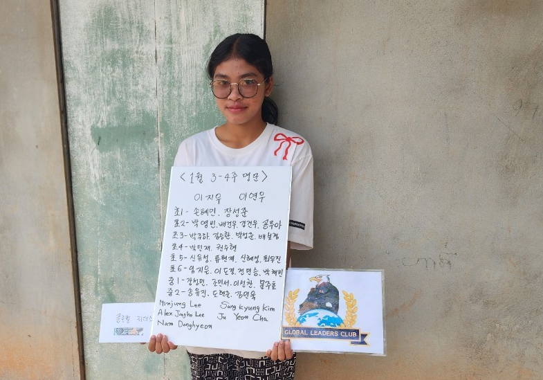 Young girl with glasses and dark hair, holding a handwritten poster and a printed sign with a logo, standing against a wall.