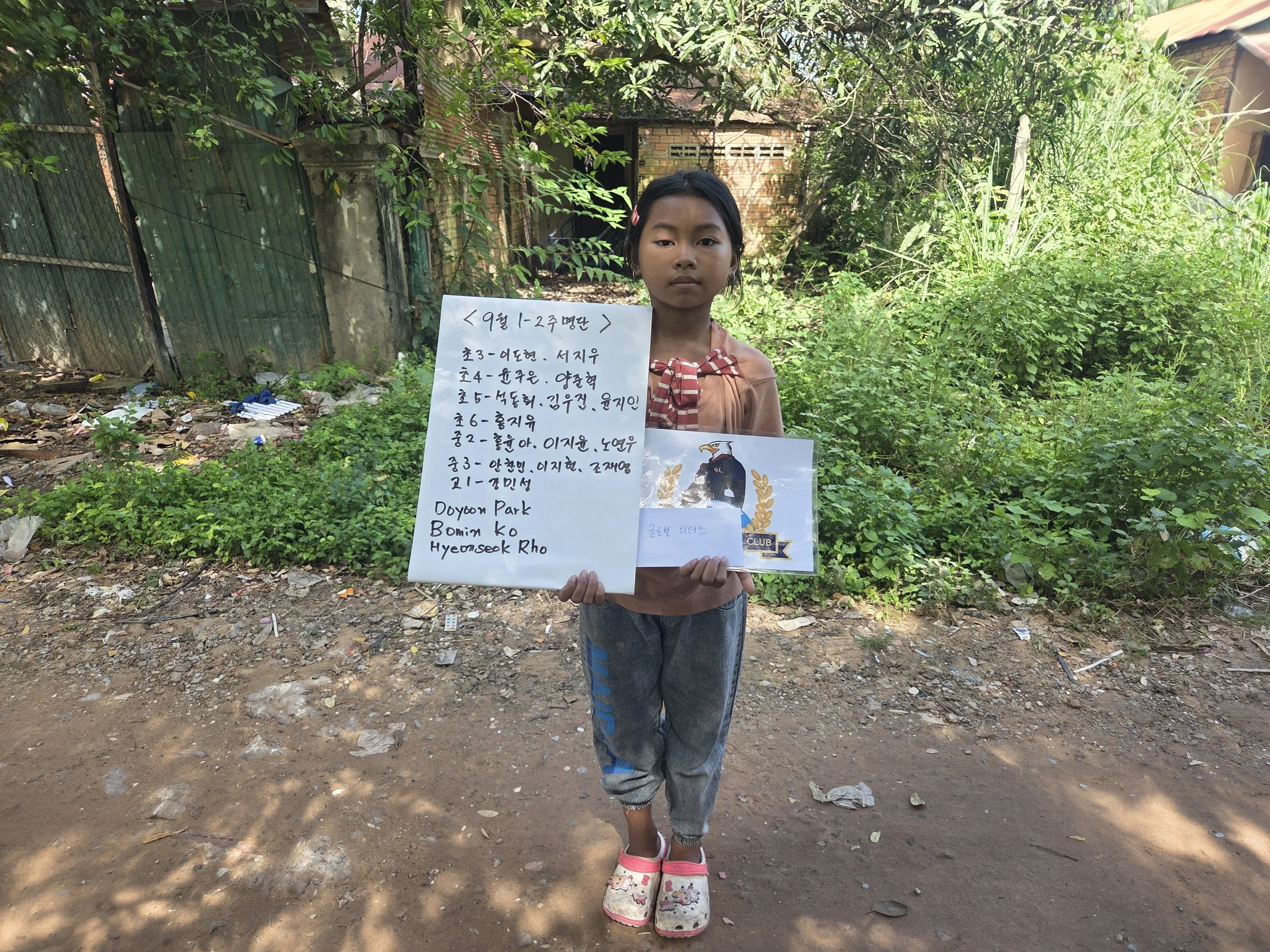 A young girl standing outdoors on a dirt path with green bushes and trees behind her, holding a whiteboard with handwritten text in Korean and some English, along with a picture of a bird and laurel leaves.