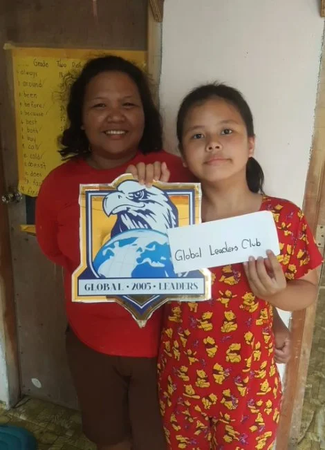 A woman and a girl standing together, holding a sign that reads "Global Leaders Club," with the woman also holding a logo featuring an eagle and the Earth, inside a school classroom.