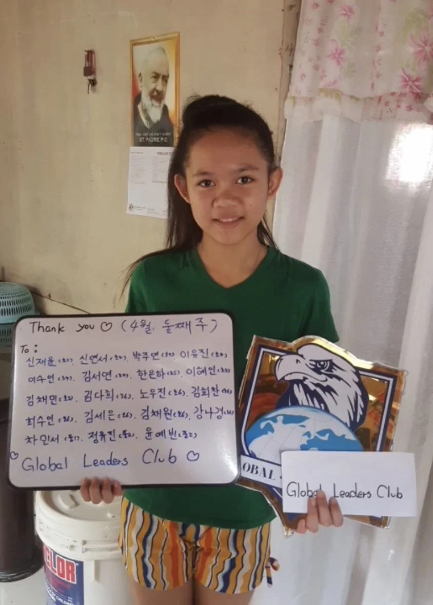 Young girl holding a sign with handwritten message and a large eagle graphic, standing indoors near a decorated wall with a framed picture of a man on it.