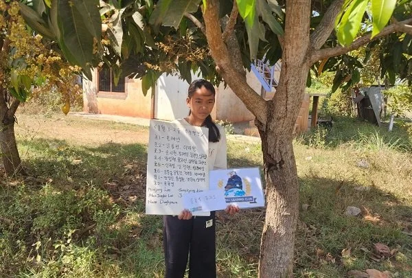 Young girl standing outdoors under a tree, holding a handwritten poster and a book.