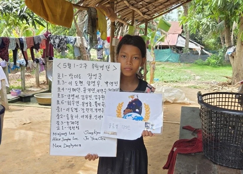 A young girl holding handwritten signs and a colorful drawing in an outdoor setting with trees and hanging laundry in the background.