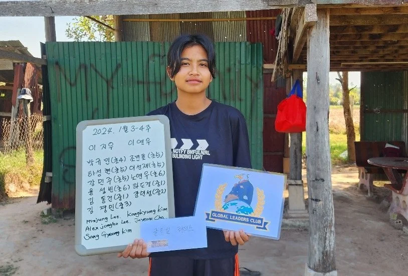 A young girl standing outdoors in front of a green corrugated metal and wooden structure, holding a whiteboard with handwritten text and a certificate with a 'Global Leaders Club' logo.