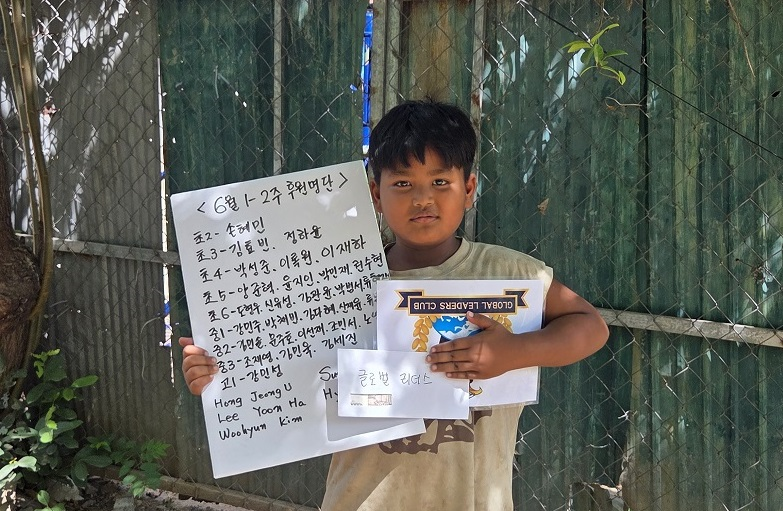 A young boy standing outdoors next to a chain-link fence, holding a book titled 'Global Leaders.' He is wearing a beige t-shirt and has dark hair. There is a whiteboard with writing in Korean and English attached to the fence behind him.