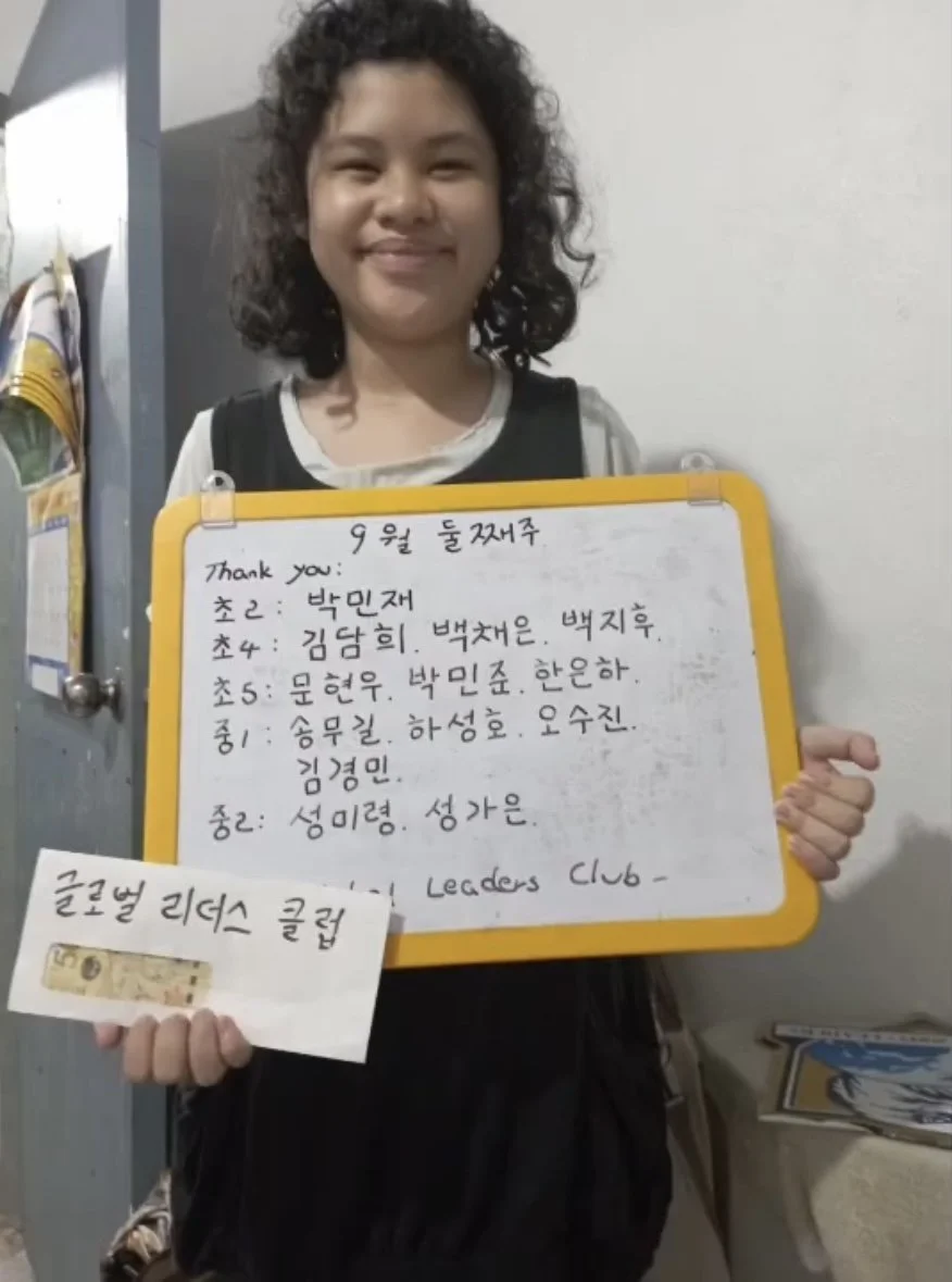 A smiling young girl with curly hair holding a whiteboard with Korean writing, standing in front of a white wall. She is wearing a black vest over a white shirt and has a backpack.