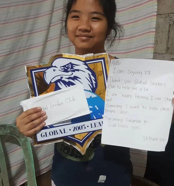 A young girl holding a letter and a large emblem of the Global Leaders Club, featuring an eagle and globe, with a striped fabric background.