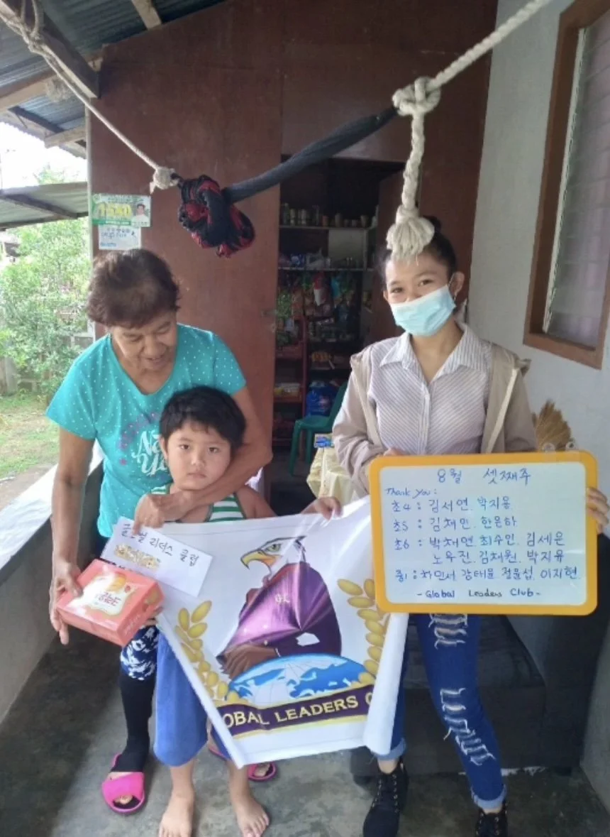 A young girl wearing a face mask holding a whiteboard with writing, standing next to a boy holding a banner, with an older woman and a smaller child in front of them. They are indoors, with some outdoor greenery visible through a window.