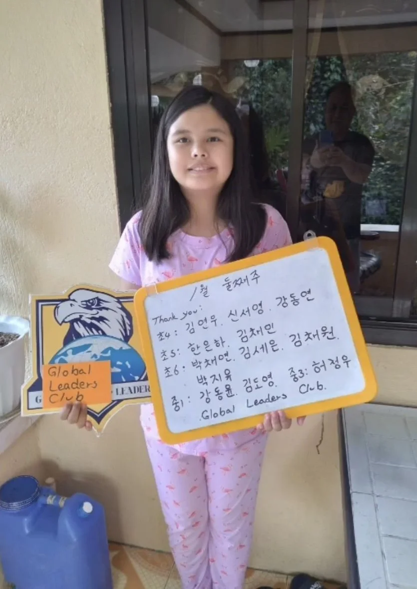 A young girl in pink pajamas standing indoors, holding a sign with handwritten notes and a logo of the Global Leaders Club. Behind her, a glass door reflects a person taking her photo.