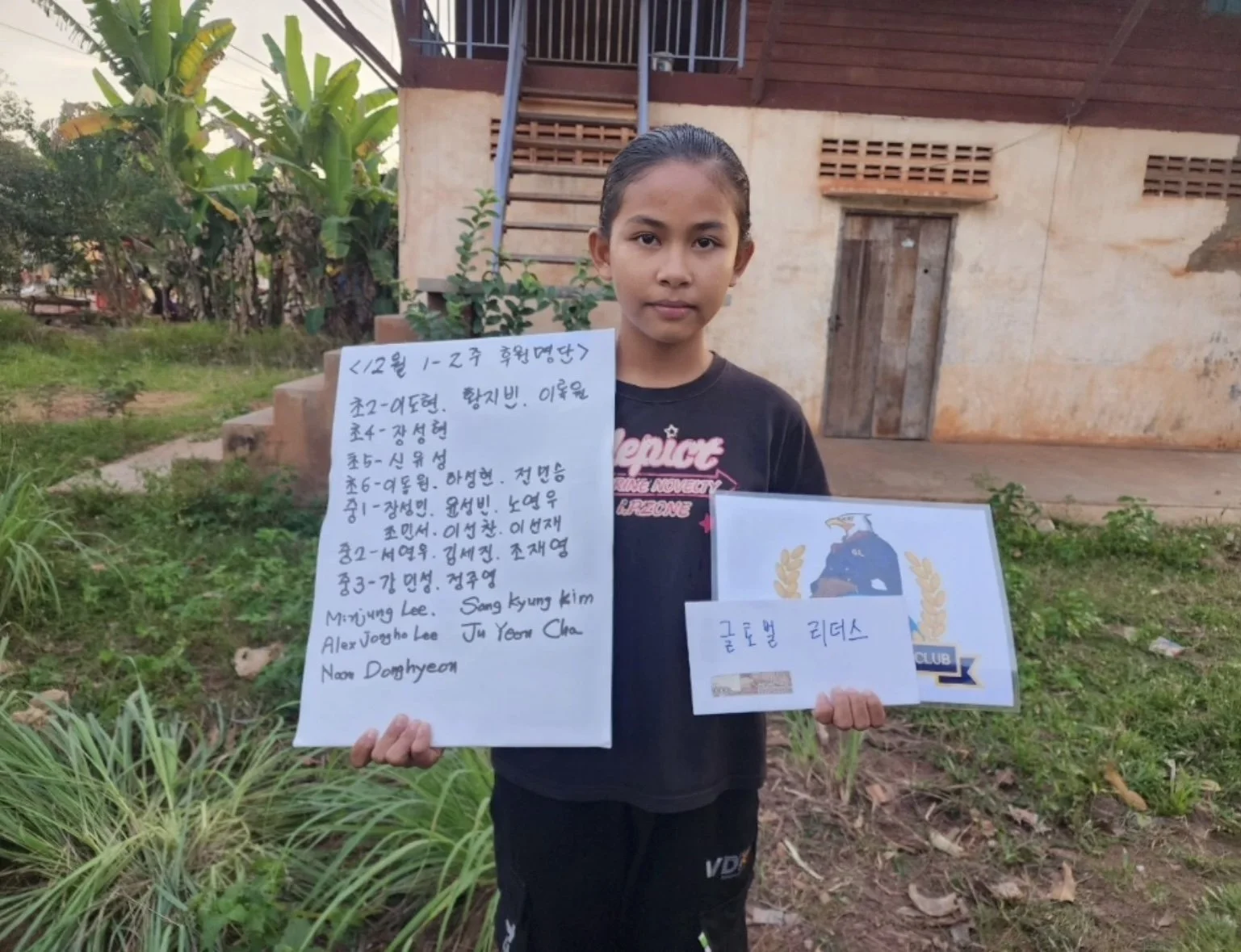 A young girl holding handwritten signs stands outdoors in front of a rustic building with a staircase and greenery. The signs contain writing in Korean and English, referencing a soccer team, the Goto Ball Ladies.