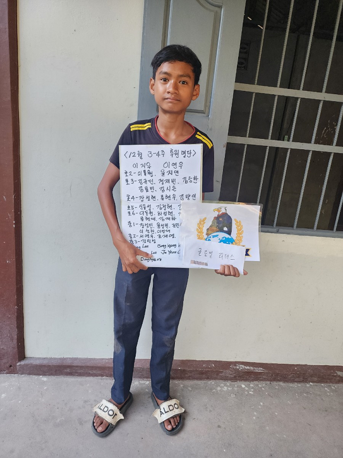A young boy stands in front of a beige wall and metal gate, holding a sign written in Korean and an illustration with a globe and a black ship. He is wearing a dark t-shirt with yellow stripes on the shoulders, jeans, and sandals labeled 'ALDO'.