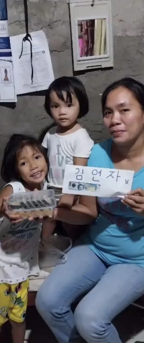 A woman and two young girls inside a room with a concrete wall. The woman is holding a handwritten sign with Korean characters and a file folder. The girl on the left is smiling and holding a plastic container with items inside, wearing yellow patter