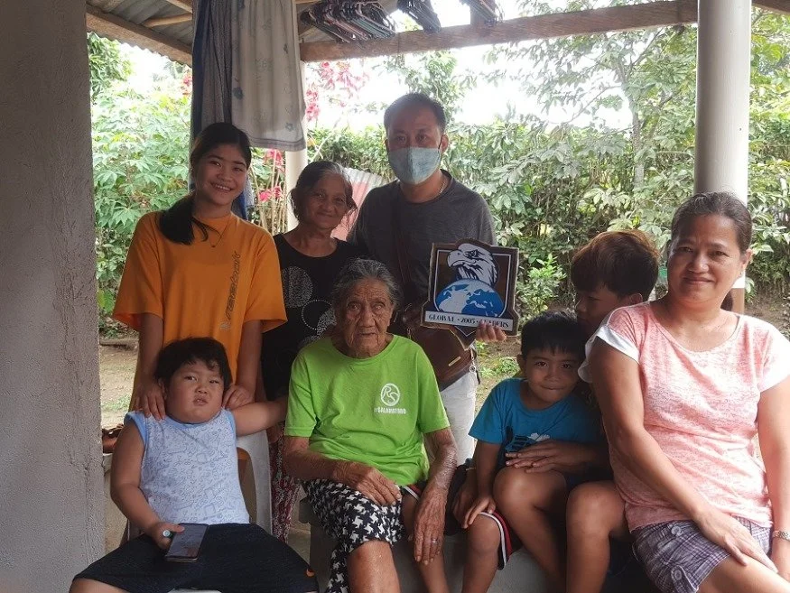 Group of seven people, including elderly woman, children, and adults, sitting and standing outside a house with green trees in the background, some smiling and one person wearing a face mask.