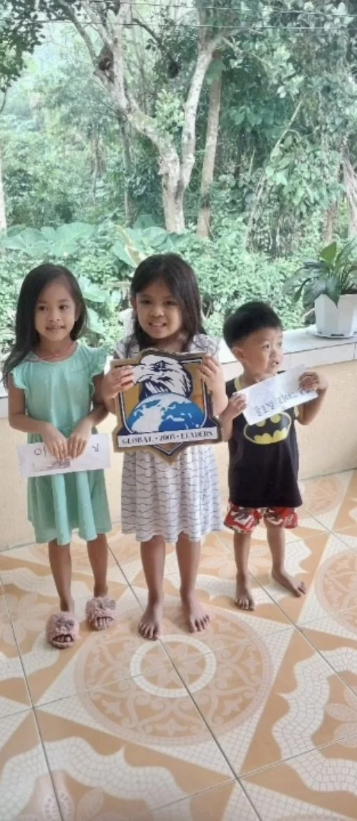 Three young children standing indoors with large windows behind them showing green trees. The girl on the left is wearing a mint green dress and slippers, holding a piece of paper. The girl in the middle is wearing a striped dress and holding a sign 