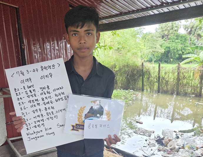 A young boy in a dark shirt holding two pieces of paper, one with handwritten text in Korean and the other with a printed image of a bird and the word 'GLOB' on it. He is standing near a pond or flooded area with trees and a fence in the background.