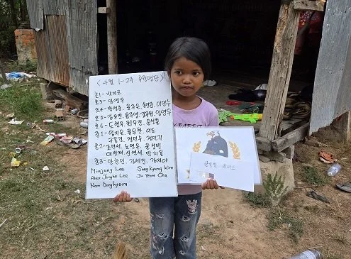 A young girl standing outdoors in front of a simple wooden structure, holding two signs with writing and illustrations. One sign has handwritten text, and the other has a printed drawing of a bear and other animals.