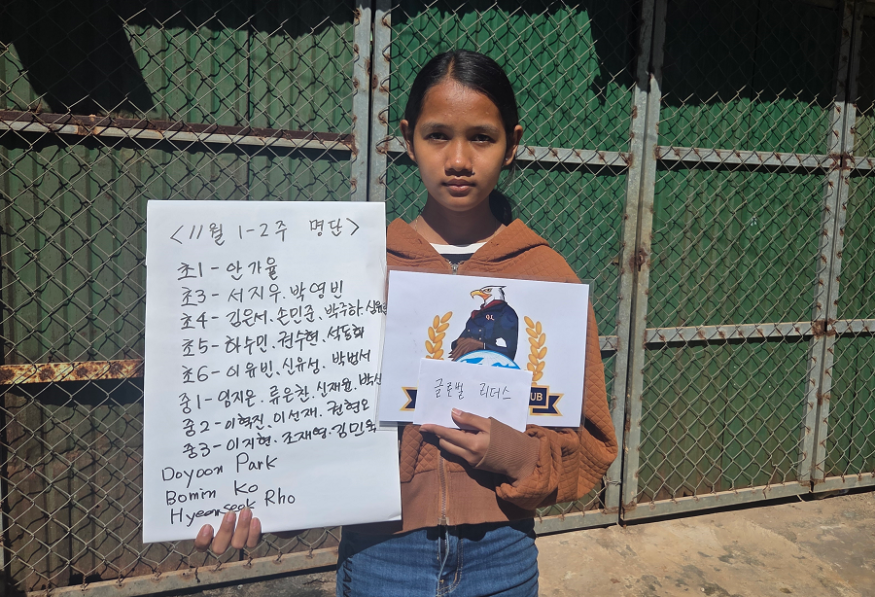 A young girl standing in front of a chain-link fence holding a handwritten list in one hand and a colorful drawing of an eagle mascot in the other. The girl is wearing a brown hoodie and has dark hair tied back.