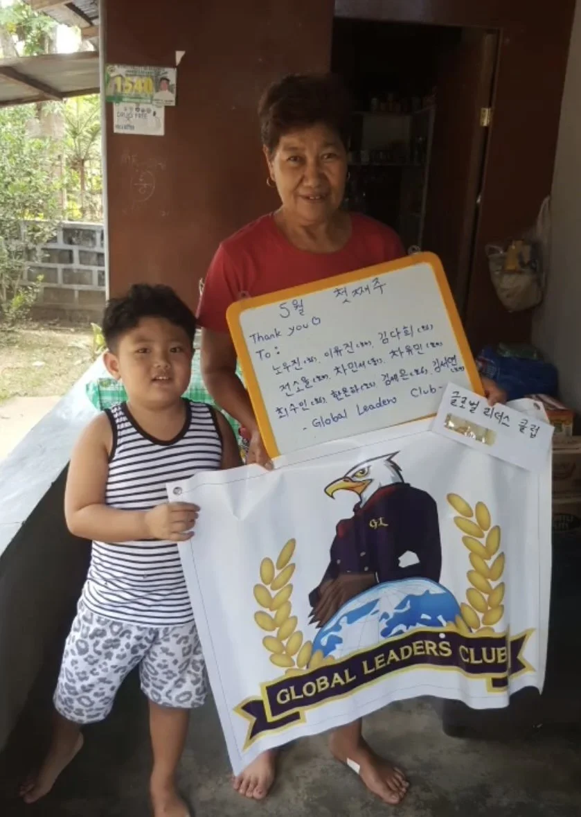 An elderly woman and a young boy standing indoors, holding a large banner of the Global Leader's Club with an eagle logo and a handwritten note.