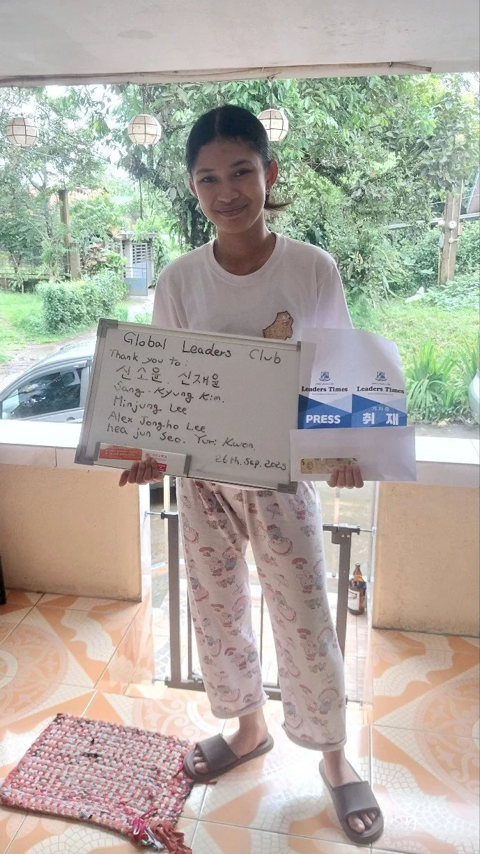 Young girl standing indoors, holding a whiteboard with handwritten messages and newspaper clippings, smiling, wearing a white t-shirt and patterned pajamas, near a window with green outdoor scenery.