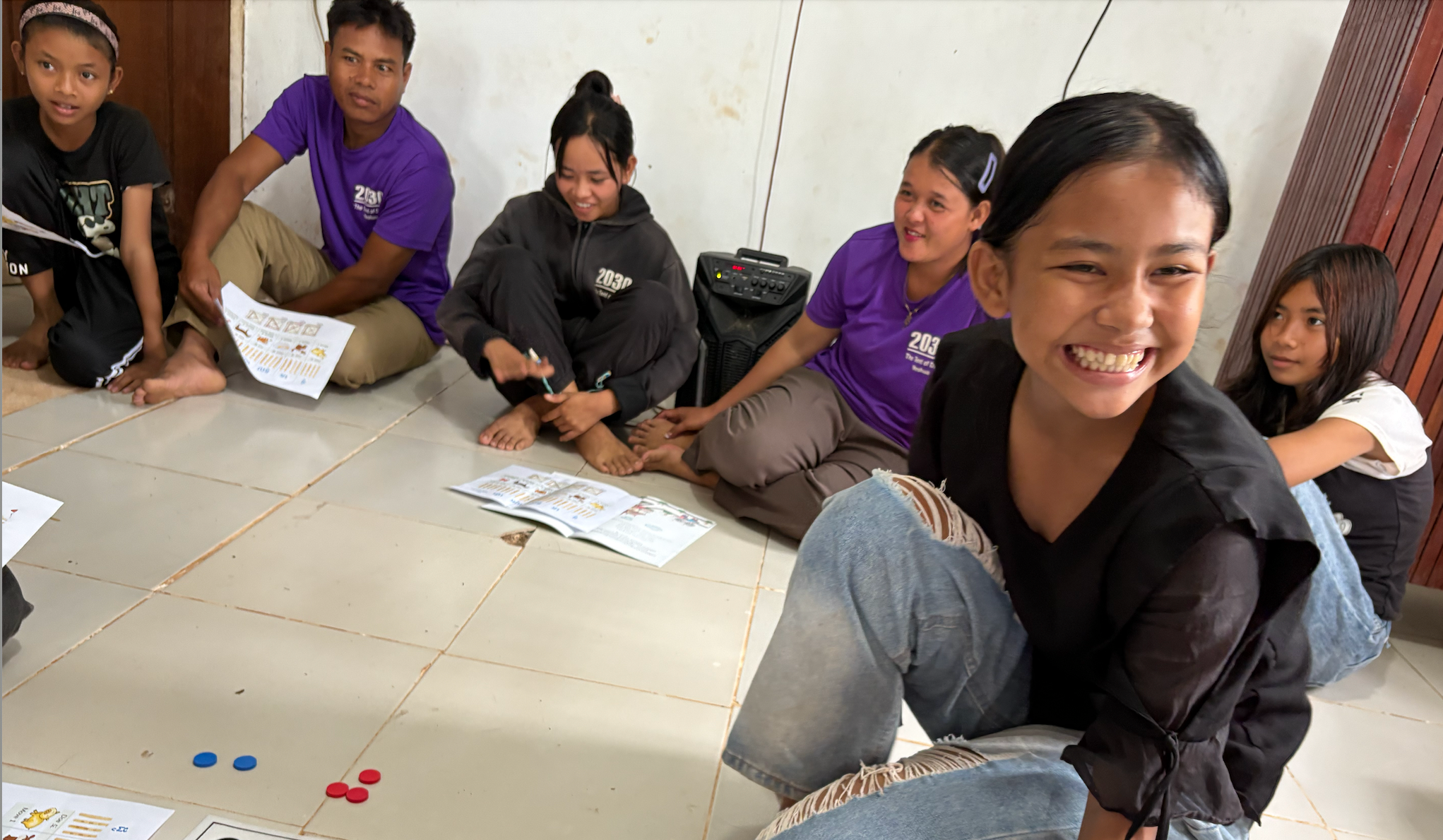 Children sitting on the floor in a circle, engaged in an activity or game, with papers and colored game pieces in front of them, and smiling or talking to each other.