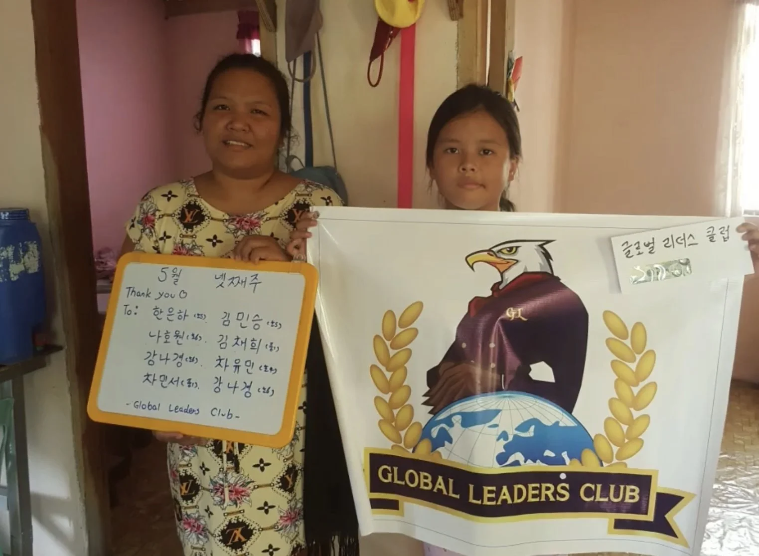 Two women standing indoors, holding a white banner with a logo of an eagle, a globe, and gold laurel leaves, representing the Global Leaders Club. One woman is holding a whiteboard with handwritten text in Korean and English, thanking members of the 