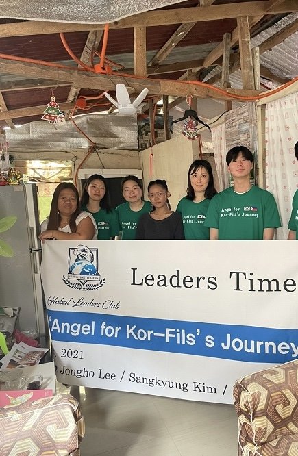 Six young women standing behind a banner that reads 'Leaders Time' and 'Angel for Kor-Fil's Journey,' inside a rustic room with wooden beams, a refrigerator, and holiday decorations.