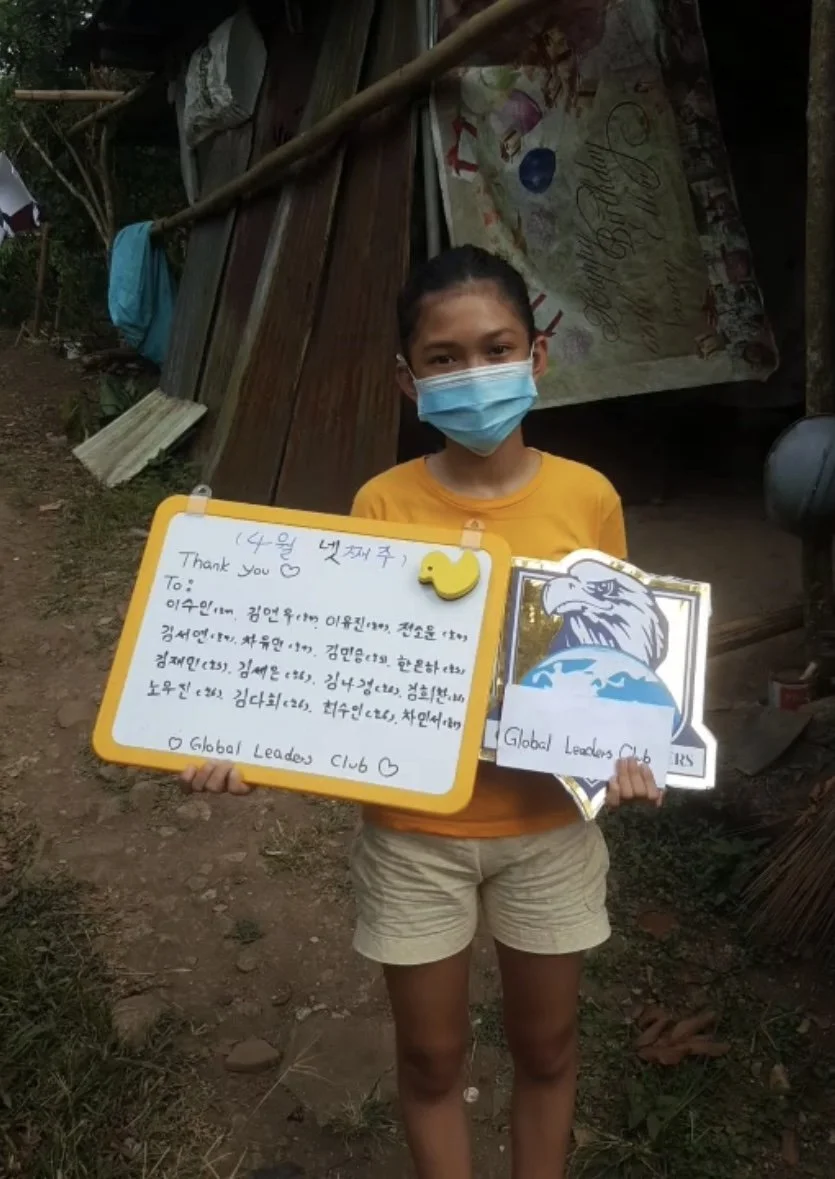 A young girl wearing a face mask and yellow shirt holding a whiteboard with handwritten message and a paper with a tiger illustration, standing outdoors in front of a wooden structure.