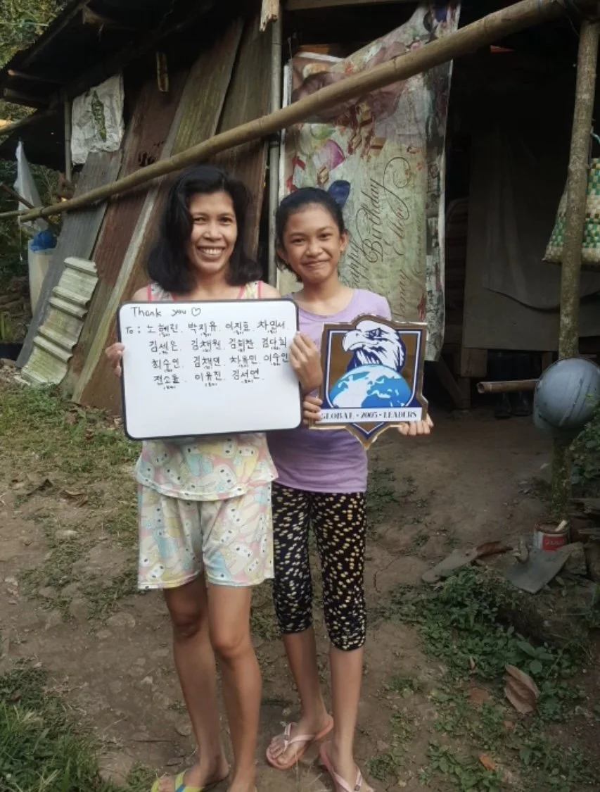 Two women, one older and one young girl, standing outdoors in front of a makeshift structure made of wooden planks, holding a handwritten note and a logo of an eagle with a globe. Both are smiling and wearing casual clothing.