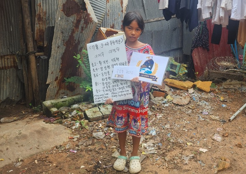 A young girl holding a cardboard sign and a printed paper standing on a dirt ground in front of a makeshift fence made of corrugated metal sheets, with clothes hanging to dry in the background.
